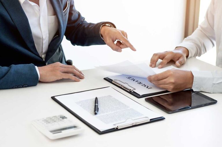 Two people reviewing car insurance documents at a kitchen table with a laptop and paperwork.